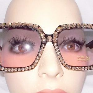 Może przedstawiać: Czarne kwadratowe okulary przeciwsłoneczne z grubą oprawką ozdobioną złotymi kryształkami. Soczewki mają gradient od ciemnofioletowego do jasnofioletowego. Okulary przeciwsłoneczne mają ochronę UV 400 i certyfikat CE.