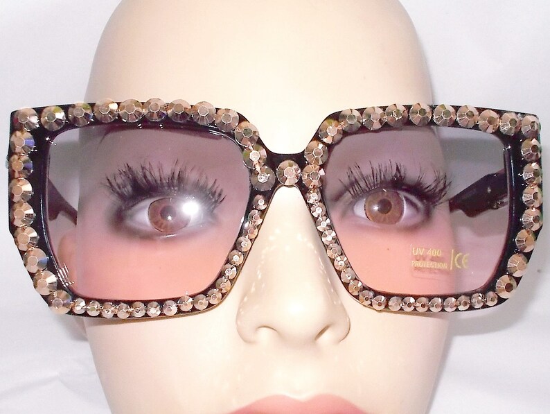 Może przedstawiać: Czarne kwadratowe okulary przeciwsłoneczne z grubą oprawką ozdobioną złotymi kryształkami. Soczewki mają gradient od ciemnofioletowego do jasnofioletowego. Okulary przeciwsłoneczne mają ochronę UV 400 i certyfikat CE.