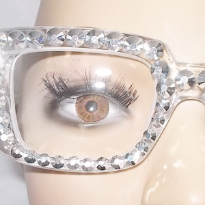 Peut inclure: Lunettes à monture en plastique transparente avec des strass autour des montures. Les lunettes sont sur le visage d'un mannequin.