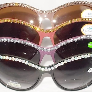 May include: Four pairs of rhinestone-embellished sunglasses with different coloured frames and lenses. The sunglasses have a cat-eye shape and are stacked on top of each other. Each pair has a sticker with the text "Sun Readers" and a magnification strength: +1.50, +2.00, +2.50, and +2.00.