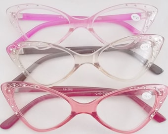 Puede incluir: Tres pares de gafas de lectura de ojo de gato rosa con diamantes de imitación alrededor de los marcos. Las gafas están apiladas una encima de la otra. El par superior es rosa, el par del medio es transparente y el par inferior es rosa oscuro. Las gafas tienen el texto "R443RS" y "+2.00" en las patillas.