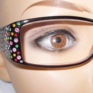 May include: Black-framed eyeglasses with clear lenses. The temples are decorated with colorful rhinestones. The glasses are on a mannequin head, showcasing the eyewear's design and style. The frames have a rectangular shape.