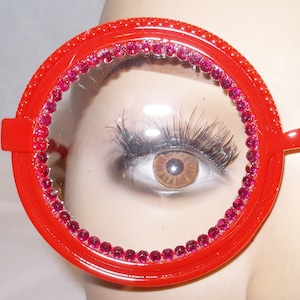 May include: Red round plastic glasses with a pink rhinestone trim around the lenses. The glasses are on a mannequin's face.