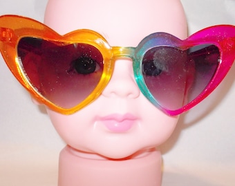 May include: Heart-shaped sunglasses with a rainbow gradient effect. The lenses are a dark brown color. The frames are made of plastic and are in shades of orange, green, and pink.