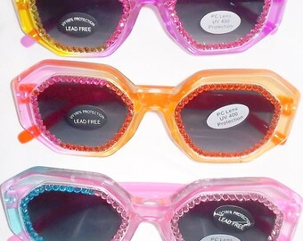 Può includere: Tre paia di occhiali da sole geometrici e colorati. Ogni paio ha una montatura di colore diverso: giallo, arancione e rosa. Le montature sono decorate con strass attorno alle lenti. Ogni lente è scura e gli occhiali da sole hanno un adesivo con scritto "UV 100% PROTECTION LEAD FREE".