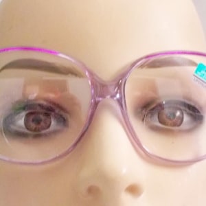 Könnte beinhalten: Eine Brille im Vintage-Stil mit einem hellvioletten Rahmen. Die Gläser sind klar, und auf einem ist ein kleiner Aufkleber zu sehen. Die Brille ist vor einem neutralen Hintergrund platziert.