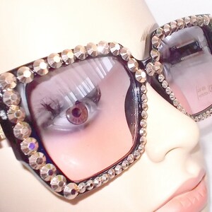 Może przedstawiać: Czarne kwadratowe okulary przeciwsłoneczne z kryształkami wok&oacute;ł oprawki i r&oacute;żowymi szkłami. Okulary mają złoty akcent z boku. Tekst "UV 400 CE" jest nadrukowany na prawym szkle.