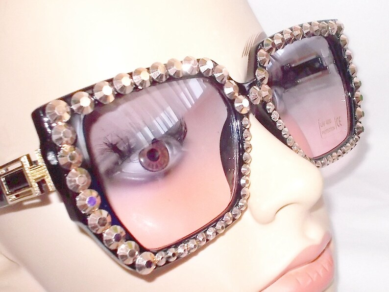 Może przedstawiać: Czarne kwadratowe okulary przeciwsłoneczne z kryształkami wok&oacute;ł oprawki i r&oacute;żowymi szkłami. Okulary mają złoty akcent z boku. Tekst "UV 400 CE" jest nadrukowany na prawym szkle.