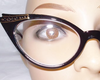 Könnte beinhalten: Schwarze Cat-Eye-Brille mit klaren Gläsern. Die Fassungen haben eine dekorative goldfarbene Verzierung an den oberen Außenecken. Die Brille ist vor einem neutralen Hintergrund platziert.