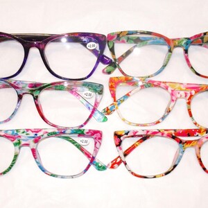 May include: Six pairs of cat-eye reading glasses arranged in two rows. The top left pair is purple, while the other five pairs feature various colorful floral and abstract patterns. Each lens has a small white sticker with the text +2.00 printed on it.