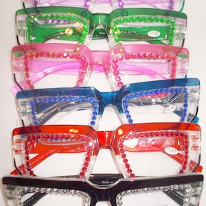 May include: Five pairs of reading glasses with rhinestones around the frames. The glasses are stacked on top of each other, with the strongest prescription on top. The top pair is pink, the next is green, then pink, blue, red, and black. The lenses are clear and the frames are made of plastic. The glasses have different strengths, ranging from +1.50 to +3.00.