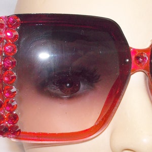 May include: Pink oversized sunglasses with a row of red rhinestones on the side. The sunglasses have a gradient lens that fades from dark to light.