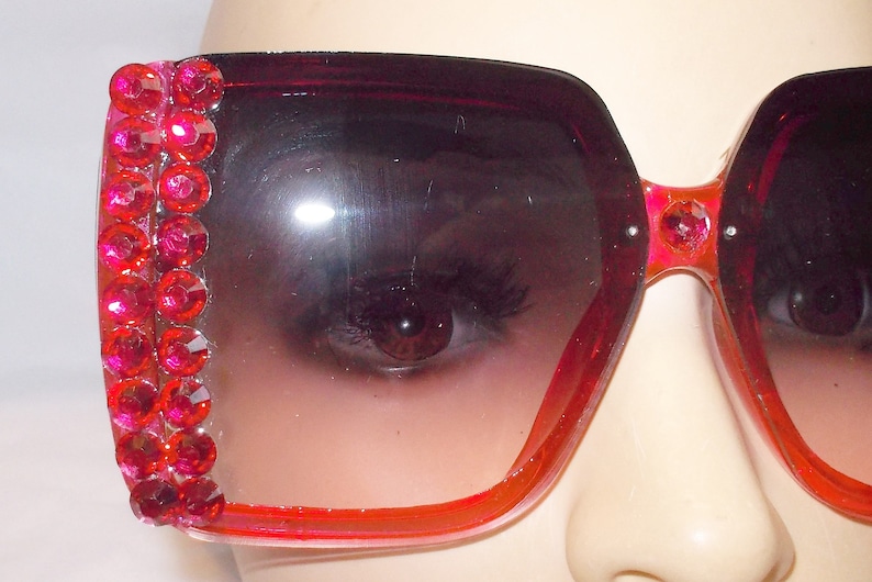 May include: Pink oversized sunglasses with a row of red rhinestones on the side. The sunglasses have a gradient lens that fades from dark to light.