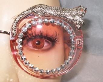 May include: Pink round sunglasses with a rhinestone trim and a silver cat design on the top of the frame.