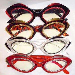 May include: Four pairs of cat-eye glasses with rhinestones around the frames. The glasses are stacked on top of each other. The top pair is red, the second pair is brown with a leopard print, the third pair is clear, and the bottom pair is red.