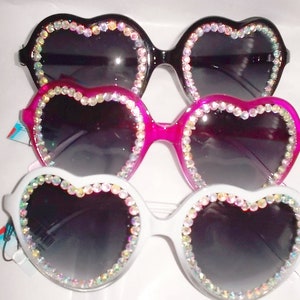 May include: Three pairs of heart-shaped sunglasses in black, pink, and white. Each has a rhinestone border and dark lenses. A stylish accessory for a fun, fashionable look.
