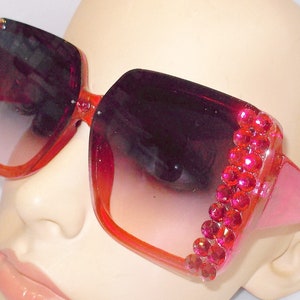 May include: Pink oversized sunglasses with a square frame and red rhinestone accents. The sunglasses have a gradient lens that transitions from dark to light.