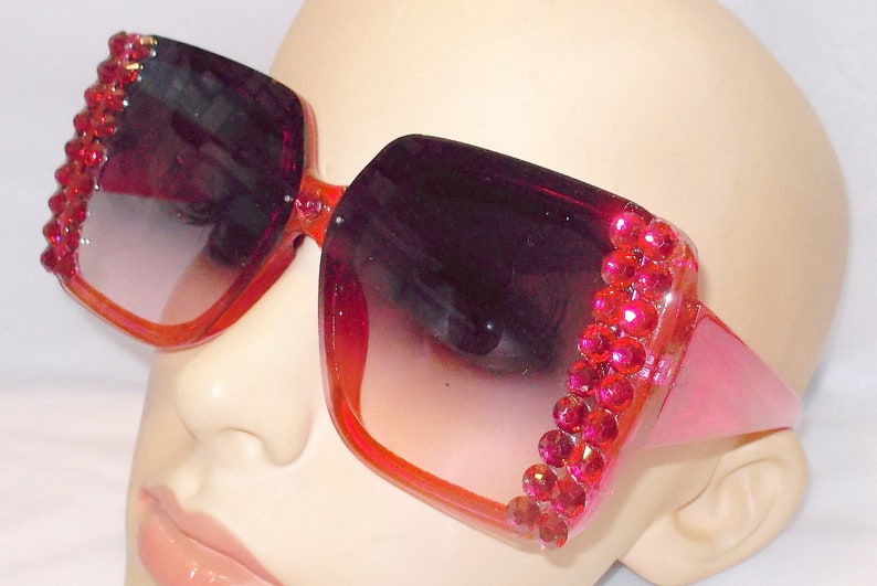 May include: Pink oversized sunglasses with a square frame and red rhinestone accents. The sunglasses have a gradient lens that transitions from dark to light.