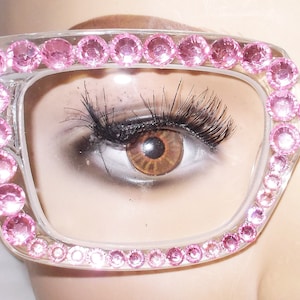 May include: Clear-framed eyeglasses with pink rhinestones outlining the frames. The glasses are square-shaped and have a glamorous, embellished design. The image shows a close-up of the glasses, with an eye visible through the lens.