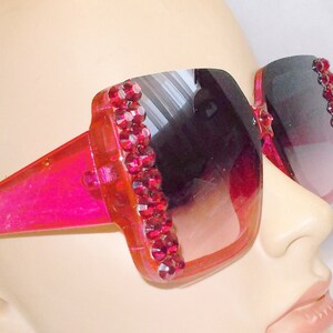 May include: Pink plastic sunglasses with a square frame and a gradient lens. The frame is decorated with a row of red rhinestones.
