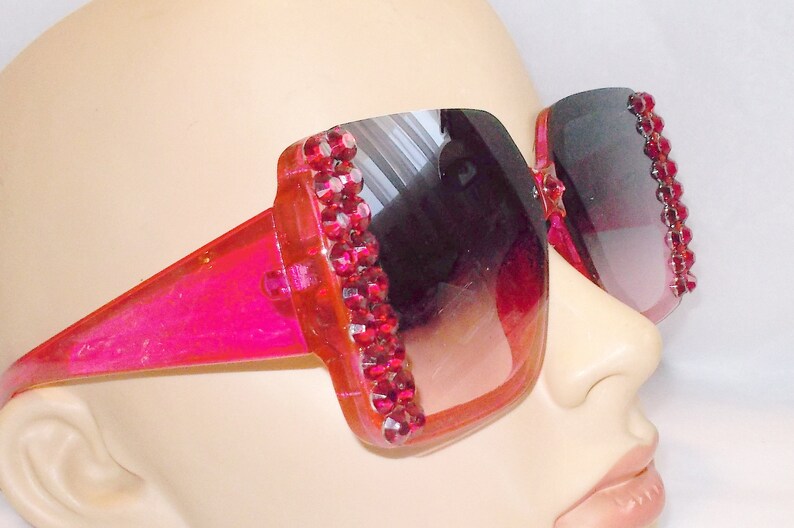 May include: Pink plastic sunglasses with a square frame and a gradient lens. The frame is decorated with a row of red rhinestones.
