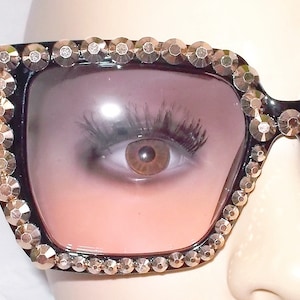 Może przedstawiać: Czarne okulary przeciwsłoneczne z kwadratową oprawą i r&oacute;żowymi szkłami z gradientem. Oprawa jest ozdobiona złotymi kryształkami.