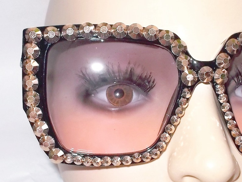 Może przedstawiać: Czarne okulary przeciwsłoneczne z kwadratową oprawą i r&oacute;żowymi szkłami z gradientem. Oprawa jest ozdobiona złotymi kryształkami.