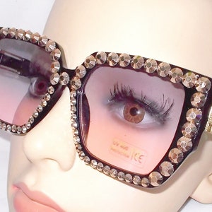 Może przedstawiać: Czarne kwadratowe okulary przeciwsłoneczne z grubą czarną oprawką i kryształkami wok&oacute;ł krawędzi. Soczewki są w kolorze przechodzącym od r&oacute;żowego do przezroczystego.