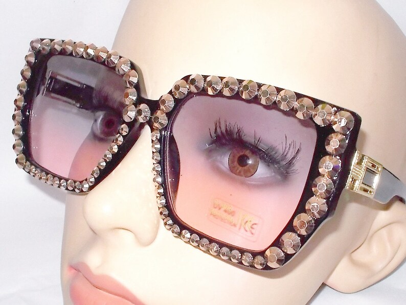 Może przedstawiać: Czarne kwadratowe okulary przeciwsłoneczne z grubą czarną oprawką i kryształkami wok&oacute;ł krawędzi. Soczewki są w kolorze przechodzącym od r&oacute;żowego do przezroczystego.