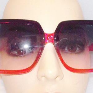 May include: A pair of oversized, square-shaped sunglasses with a pink frame and pink rhinestone accents. The lenses are a gradient of dark pink to light pink.