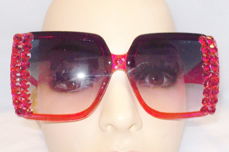 May include: A pair of oversized, square-shaped sunglasses with a pink frame and pink rhinestone accents. The lenses are a gradient of dark pink to light pink.