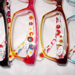 Puede incluir: Cuatro pares de gafas con monturas coloridas. Las monturas son negras, rosas, marrones y azules. Las patillas de las gafas tienen una base blanca con lunares coloridos. Cada par tiene una etiqueta de +3,50.