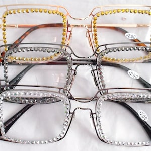 Könnte beinhalten: Drei Brillen mit quadratischem Rahmen und dekorativen Strasssteinen. Die Rahmen sind goldfarben, silberfarben und schwarz. Jede Brille hat klare Gläser und einen +2,00 Aufkleber auf dem Glas.