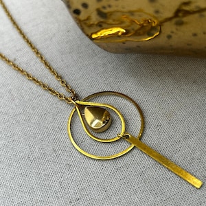 May include: A gold-colored necklace with a delicate chain. The pendant features a teardrop shape with a gold bead inside a gold ring, and a rectangular bar. The necklace is displayed on a textured, light gray surface.