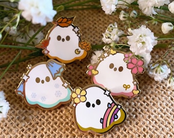 Könnte beinhalten: Vier Emaille-Pins mit niedlichen Geisterdesigns in verschiedenen saisonalen Themen. Die Geister sind weiß mit schwarzen Augen und mit verschiedenen Farben und Details verziert. Die Pins sind goldfarben und haben eine glänzende Oberfläche.