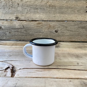 Pode incluir: Uma caneca de esmalte branco com uma borda preta. A caneca está sobre uma superfície de madeira.