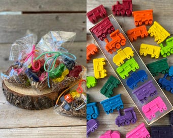 May include: A collection of colorful, train-shaped crayons in various colors, including red, orange, yellow, green, blue, purple, and pink. Some crayons are packaged in clear cellophane bags tied with colorful twine, and others are displayed in a box.