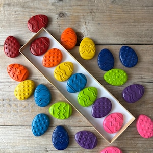 Puede incluir: Una colección de ceras en forma de huevo de colores brillantes, incluyendo rojo, naranja, amarillo, azul, verde y morado. Cada cera presenta un diseño texturizado único. Algunas ceras están dispuestas en una caja rectangular, otras dispersas.