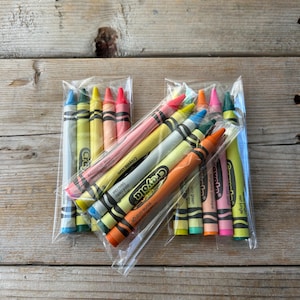 May include: Several packs of Crayola crayons in various colors, including blue, green, yellow, orange, and pink. The crayons are in clear plastic packaging and are arranged on a wooden surface. The text "Crayola" is visible on the crayons.