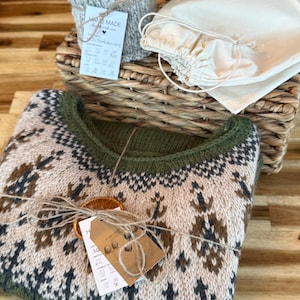 May include: A folded knit sweater featuring a cream, brown, and dark green geometric pattern with a ribbed olive green neckline. The sweater is tied with twine and a dried orange slice, accompanied by a tag, a rolled knit item, and a drawstring bag.
