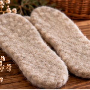 May include: A pair of beige, textured felted wool insoles with a rounded toe and heel shape. The surface of the insoles features a dense, fibrous, and slightly mottled appearance with a soft, fuzzy pile, designed to fit inside footwear.