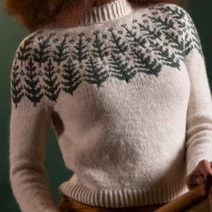 May include: A cream-colored long-sleeve sweater featuring a ribbed mock neck and a dark green knit pattern of stylized pine trees encircling the yoke and shoulders. The garment has a ribbed hem and a soft, textured knit construction throughout.