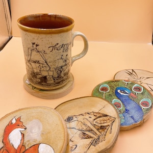 May include: Set of five wooden coasters with various designs. One coaster features a fox, another a frog, a third a peacock, and two coasters are plain wood. The coasters are all round and have a natural wood finish.
