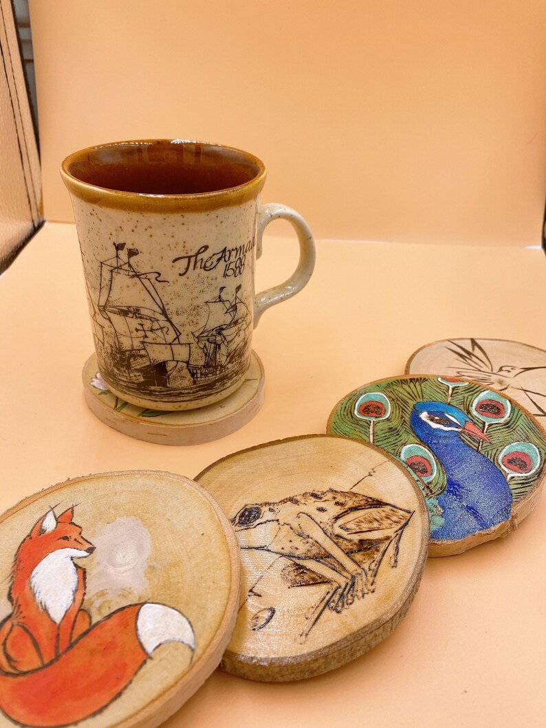 May include: Set of five wooden coasters with various designs. One coaster features a fox, another a frog, a third a peacock, and two coasters are plain wood. The coasters are all round and have a natural wood finish.
