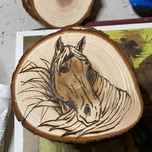May include: A wood slice with a woodburned image of a horse's head. The horse is facing the viewer and has a brown coat. The wood slice is a light brown color and has a natural wood grain.