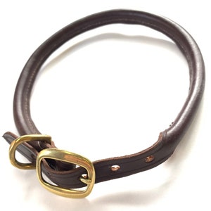 May include: A brown leather dog collar with a brass buckle. The collar is lying on a white surface.
