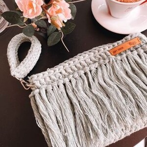 May include: A light gray crocheted bag with a long fringe. The bag has a brown leather tag with the text "Drift" on it. The bag is sitting on a table with a cup of coffee and flowers.