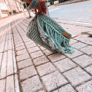 May include: A light green crocheted bag with fringe detailing and a brown leather tag that says "Soft Touch".