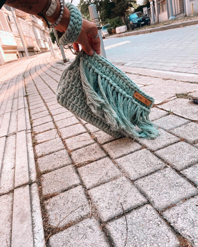 May include: A light green crocheted bag with fringe detailing and a brown leather tag that says "Soft Touch".