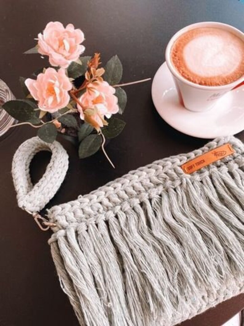 May include: A light gray crocheted bag with a long fringe. The bag has a brown leather tag with the text "Drift" on it. The bag is sitting on a table with a cup of coffee and flowers.